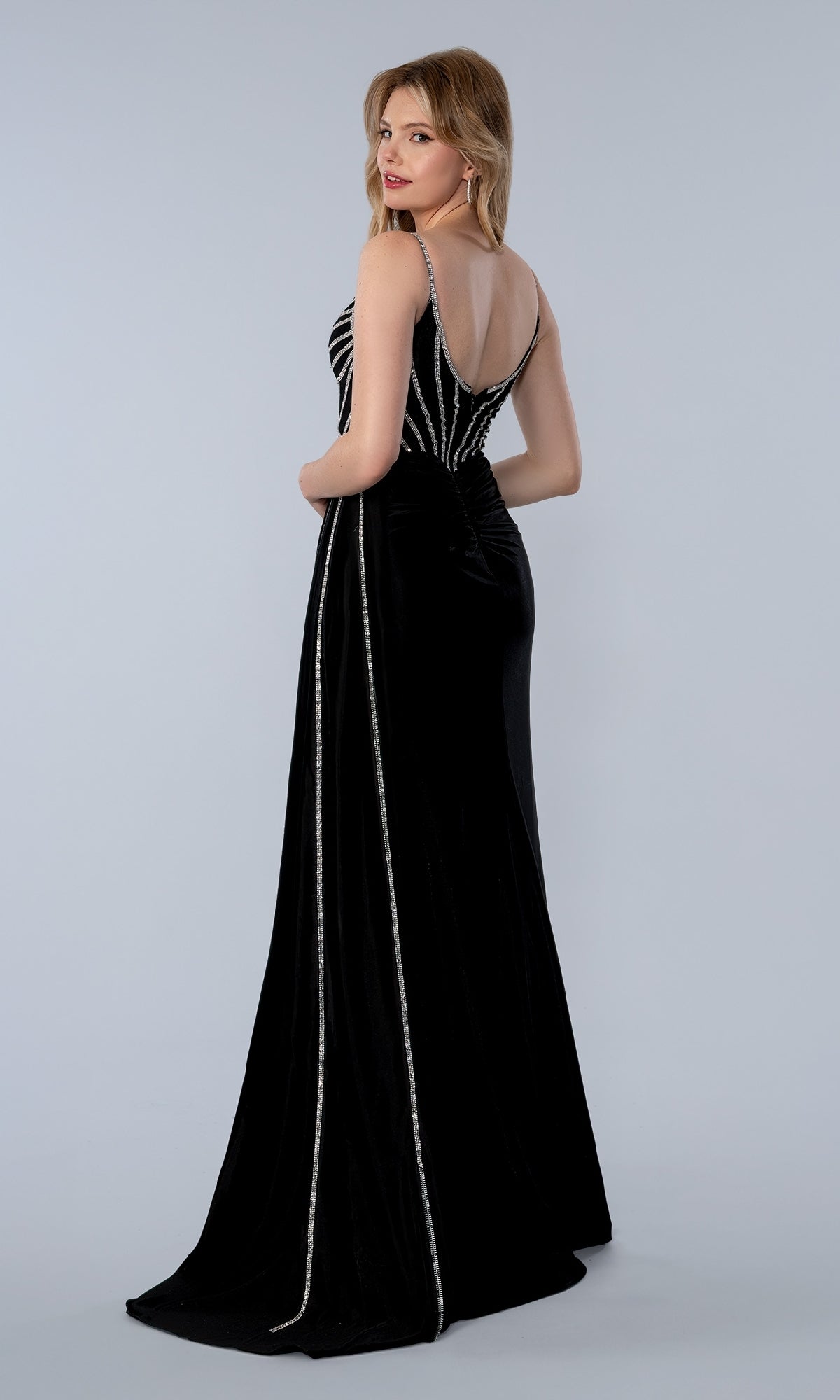 A woman stands facing backward in the black Long Prom Dress Stella Couture 26166, adorned with silver rhinestone embellishments, against a plain light gray background.
